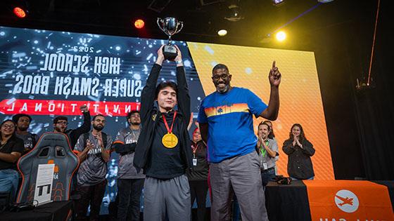 Two people are standing side by side on a stage while one holds a large silver trophy over their head.